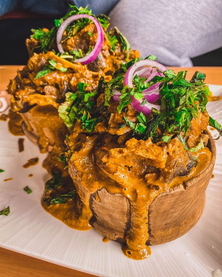 Traditional Nigerian Nkwobi served in a wooden bowl, featuring tender cow foot in spicy palm oil sauce, garnished with sliced red onions and fresh utazi leaves